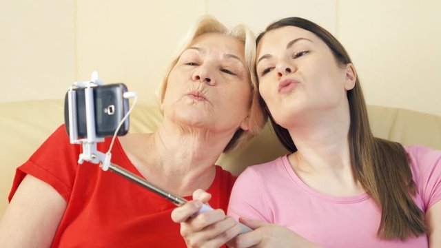 Mother and daughter sitting on couch at home. Making selfie with smartphone and selfie stick. Family making silly faces having fun. Active modern lifestyle of older people