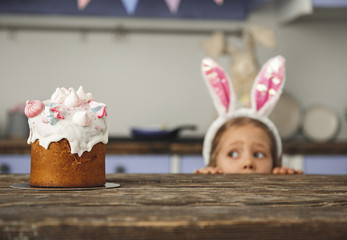 Cute little sweet tooth in bunny ears headband peeking out from behind kitchen table and looking at tasty easter cake with interest. Focus on pastry