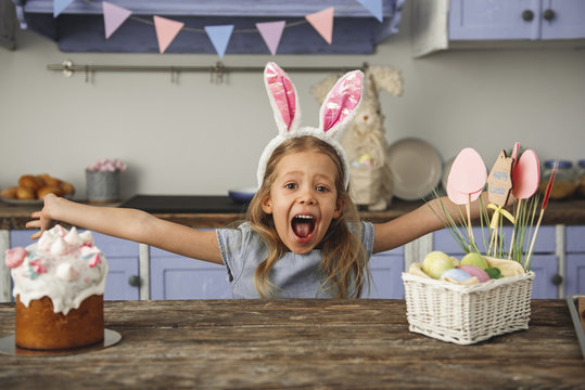 Portrait of cheery little girl in bunny ears headband standing at kitchen table with easter cake and basket with eggs and screaming with outstretched arms