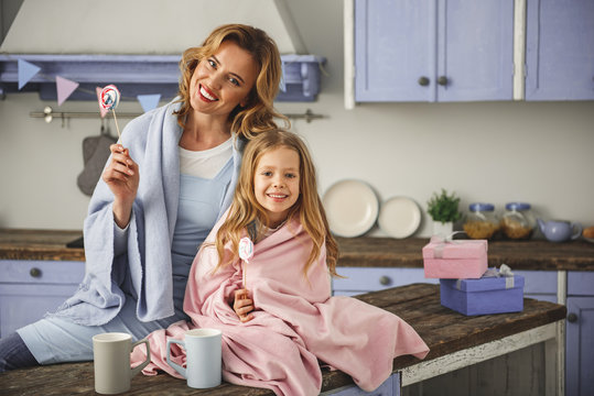 Portrait of laughing mother and daughter sitting in the kitchen with lollipops in hands. Boxes with gifts and cups are on the table. Copy space in right side