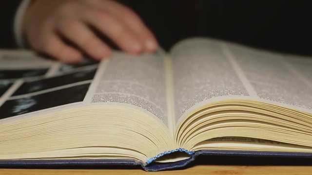 A person turns over pages of a thick illustrated encyclopedia. The businessman is looking for information in the library under the light of a desk lamp.