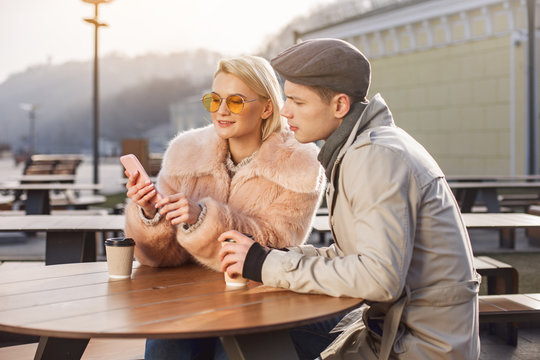 Look here. Involved young boyfriend and girlfriend are sitting at table in open air restaurant and drinking hot coffee while using modern smartphone. They are looking at screen of gadget with interest