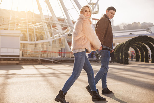 Just be happy. Full length of positive young couple are walking outdoors together. They are holding hands and looking at camera with smile. Copy space in the left side