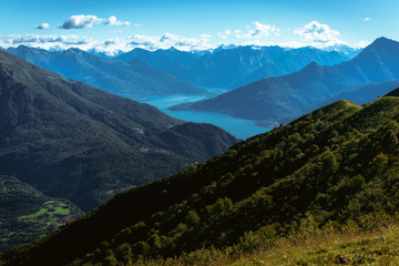 Fototapeta premium Panoramic View of beautiful landscape in the Italian Alps with fresh green meadows and snow-capped mountain tops in the background on a sunny day with blue sky and clouds in springtime.