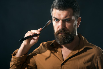 Handsome bearded hairdresser in stylish clothes holding a dangerous razor. Bearded attractive macho with long beard and moustache, holds straight razor near face. Copy space for advertise barbershop.