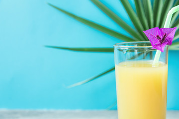 Tall Glass with Freshly Pressed Pineapple Orange Coconut Juice Straw and Small Flower. Round Palm Tree Leaf on Blue Background. Vacation Summer Relaxation Travel Healthy Superfoods Concept.