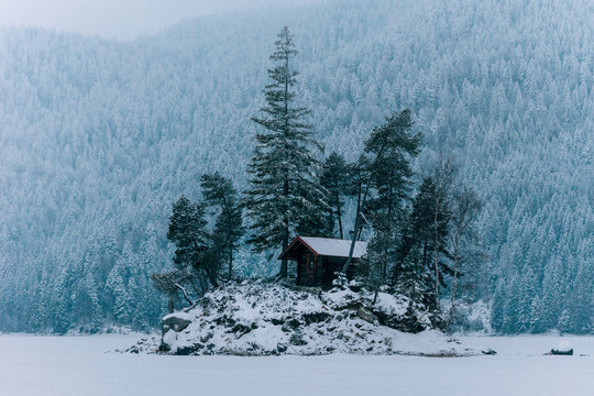  Cabin on an island on a frozen lake with trees and snowy pine forest in the background on a cloudy day at lake Eibsee, Germany