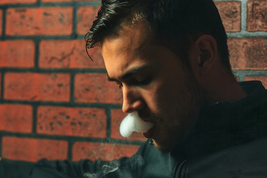 Vape man. Indoor portrait of a young handsome white guy letting of steam from an electronic cigarette. Vaping process. Close up.