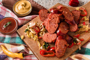 Sliced sausage and mix of fresh vegetables served on a rustic wooden board, mustard, ketchup, fresh hot chili peppers and herbs.