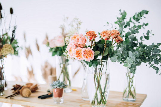 close-up of pink roses in a glass vase on a wooden table on a background of roses, dried cones, greens, scissors, rope, bumps on a white wall background