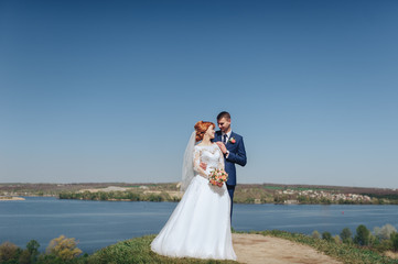 Beautiful young bride and groom stand on a cliff in the background of the river and gently embrace each other. The red-haired bride.