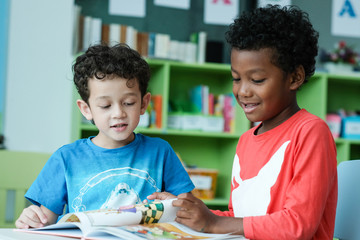 American and African boys are reading together with happiness in their kindergarten classroom, kid...