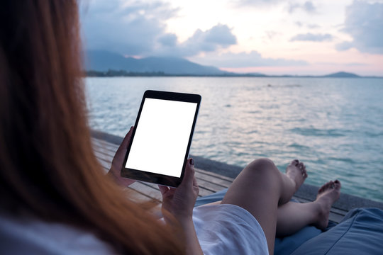 Mockup image of a woman holding and using black tablet with blank white desktop screen while sitting by the sea with blue sky background