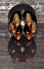 A speed loader for a revolver, for either a 357 magnum or a 38 special. It is fully loaded with full metal jacket bullets with a wooden background and showing it's reflection on the table it's on.