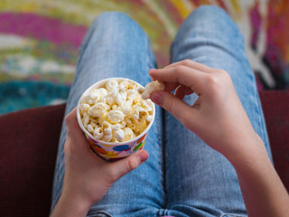 The girl eats popcorn from a colorful paper Cup sitting in a red chair.