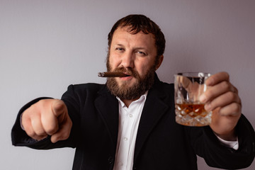 Bearded man in modern coat and shirt smoking his cigar and drink some strong liquor pointing with finger.