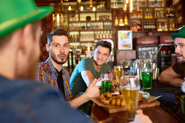 Group of young men chatting animatedly with each other and drinking alcoholic beverages while hanging out in modern pub, some of them wearing green hats