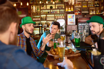 Group of handsome young friends enjoying each others company in modern pub: they drinking beer, eating snacks and chatting with each other, one of them and bearded barman wearing green hats