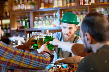 Cheerful young men toasting with beer glasses while hanging out in modern pub, handsome bearded...