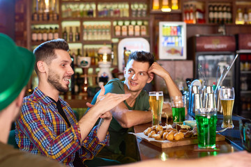 Profile view of smiling bearded man wearing checked shirt sitting at bar counter and telling funny story to his friends, they listening to him with interest, interior of modern pub on background