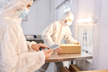 Joint work in pharmaceutical laboratory: unrecognizable worker wearing safety mask and coverall standing at lab bench and packing medicinal products while his superior keeping eye on him