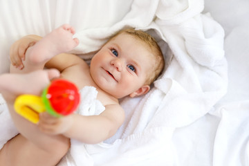 Cute little baby playing with toy rattle and own feet after taking bath. Adorable beautiful girl wrapped in white towels