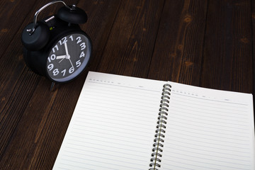 Alarm clock and notebook with pen on dark wooden floor, empty space for text.