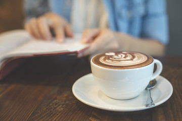Closeup of a coffee cup in the cafe add the filter retro color tone. Hand of woman reading book at coffee shop.