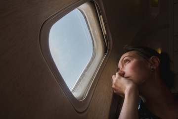 Sad girl at the porthole in the plane. Young woman on passenger seat near window in airplane