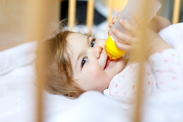 Cute little baby girl holding bottle with formula mild and drinking. Child in baby cot bed before...