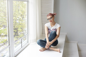 People, lifestyle, leisure and relaxation concept. Picture of stylish young pink haired woman wearing trendy eyeglasses, choker and jeans sitting barefooted by large window, enjoying good day