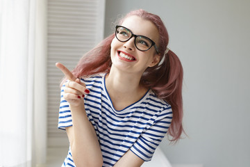 Cute cheerful female teenager in spectacles sitting in light bedroom, smiling happily and pointing finger sideways as she sees something exciting. Human facial expressions and body language