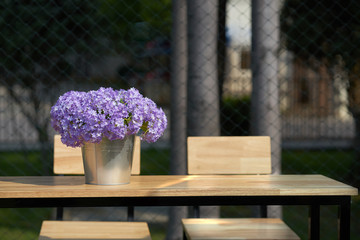 purple flowers in bucket vase on wooden table with chairs with sunlight and shade