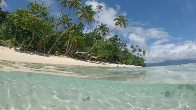 HALF UNDERWATER: Mesmerizing view of the sky above sandy shoreline from the clear blue water surrounding it. Exploring underwater world on Fiji island. Glass like sea surface glistening on ocean floor