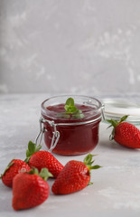 Strawberry jam in a glass jar with berries, gray background.