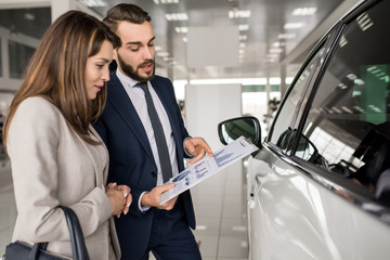 Side view portrait of modern young woman listening to car dealer while choosing luxury car in...