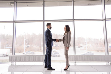 Side view full length portrait of two business partners shaking hands standing against big window in office building