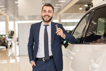 Portrait of handsome car salesman a holding car keys and smiling cheerfully at camera posing in luxury dealership showroom