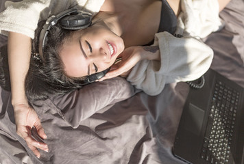Beautiful young woman lying on bed and listening music from laptop computer via headphones