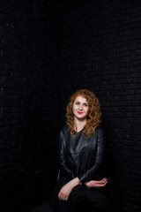 Curly hair girl at leather jacket on studio against black brick wall.