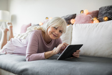 Happy senior lady lying on couch using tablet