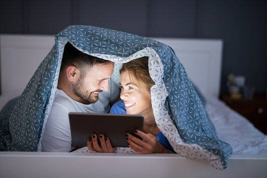 Close up view of beautiful smiling young wife and husband looking a tablet under a blanket on the bed at night. Relationship and weekend goals.