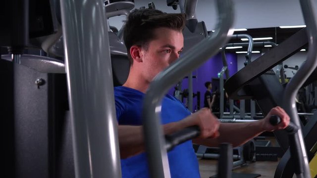 A young fit man trains on a machine in a gym - closeup from the side