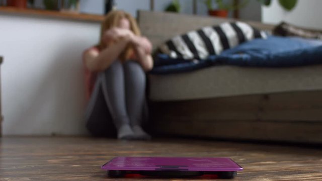 Blurry depressed woman sitting on wooden floor near bed holding head and arms on knees after weight loss fail with weight scales in foreground. Sad female upset with weight gain in domestic interior.