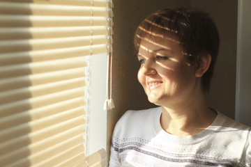 Middle-aged woman standing in front of a window in daylight, a shade of blinds on her face