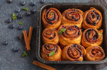 Homemade cinnamon buns with blueberries and cinnamon in oven dish on a dark background, top view, copy space.