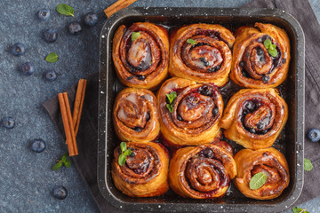 Homemade cinnamon buns with blueberries and cinnamon in oven dish on a dark background, top view, copy space.