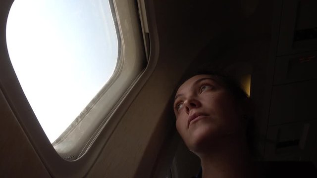 Sad girl at the porthole in the plane. Young woman on passenger seat near window in airplane