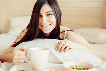 Obraz na płótnie Canvas Breakfast in bed / beautiful brunette woman holding white porcelain cup in hand and smiling lying in bed