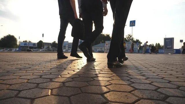 Feet of three businessmen walking near airport with sun flare at background. Business men go to terminal together. Confident guys being on his way to work trip. Colleagues going outdoor. Slow motion
